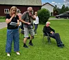 Tre personer står og sitter på en grønn plen foran røde hus og en parkert bil.