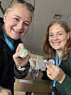 To kvinner smiler og viser frem små buttons innendørs.
