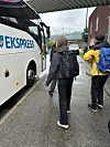 To personer går mot en hvit ekspressbuss ved et overbygd stoppested i regnvær.