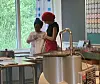 To personer står ved en kjøkkenbenk i et lyst kjøkken med stor gryte og vinduer i bakgrunnen.