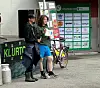 To personer står ved et sykkelstativ foran en butikkvegg med plakater og logoer.