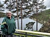 Kvinne i grønn jakke står ved et rekkverk med en fjord og trær i bakgrunnen.