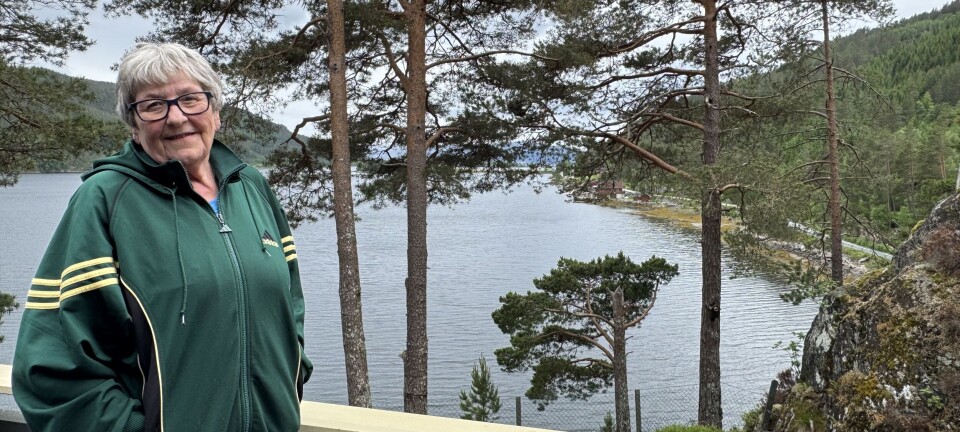 Kvinne i grønn jakke står ved et rekkverk med en fjord og trær i bakgrunnen.