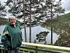 Kvinne i grønn jakke står ved et rekkverk med en fjord og trær i bakgrunnen.