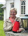 Smilende kvinne holder en rød termokanne ved et hvitt hus.