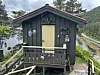 Liten, mørk trebygning med gul dør, omgitt av skog og fjell. Fjord i bakgrunnen.