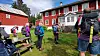 Personer med ryggsekker står samlet på et gressområde foran røde bygninger.