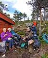 Turgåere sitter på steiner og en trestamme i skogsterreng med ryggsekker og turutstyr.
