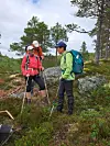Tre turgåere med sekker og staver står i et kupert skogsområde med steiner og furuer.