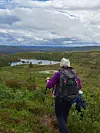 Turgåer med ryggsekk og staver på en grønn høyde med innsjø og fjellandskap under overskyet himmel.