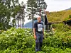 En mann står i en grønn hage med blomster og trær, med fjell i bakgrunnen.