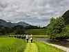 Turgåere går langs en smal sti ved elva i et grønt landskap med åser og skyer.