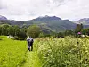 To personer går på en sti i et grønt fjellandskap med åser, trær og overskyet himmel.