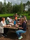 Flere personer sitter rundt et piknikbord utendørs ved vann og skog.