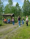 Flere personer samles ved et lite skur i en skoglysning omgitt av høye trær.