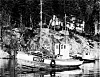 En liten båt ligger ved en steinete kyst med skog og en liten bygning på klippen.