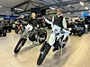 To personer på offroad-motorsykler inne i en motorsykkelutstilling med flere kjøretøy i bakgrunnen.