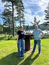 To personer står ved et utendørs ildsted i en park med gress, trær, vann og fjell.