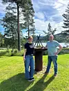 To personer står ved et utendørs ildsted i en park med gress, trær, vann og fjell.