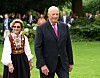 Det norske kongeparet går i en hage med grønn plen og blomster i bakgrunnen.