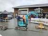 En blå minigraver står på et vått asfaltert område ved en bygning, med flere personer og maskiner i bakgrunnen.