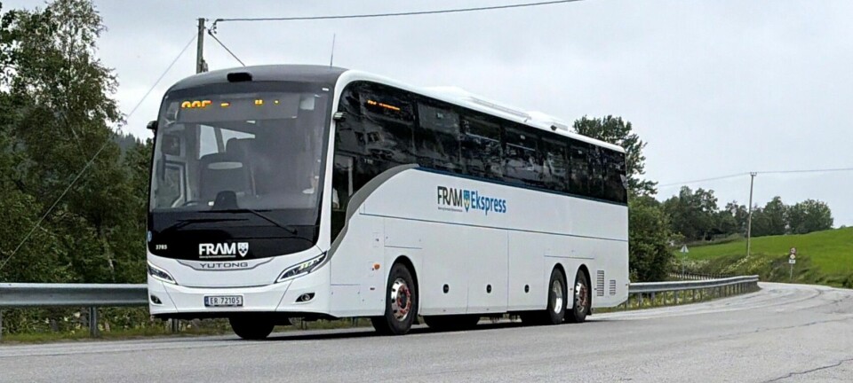 Hvit buss ved en svingete vei med gress, trær og et busskur i bakgrunnen.