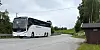 Hvit buss ved en svingete vei med gress, trær og et busskur i bakgrunnen.