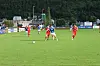 Fotballspillere i rødt og blått kjemper om ballen på en grønn bane med tribuner og skog i bakgrunnen.