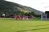 Fotballspillere i røde og hvite drakter foran mål på en gressbane med fjell i bakgrunnen.