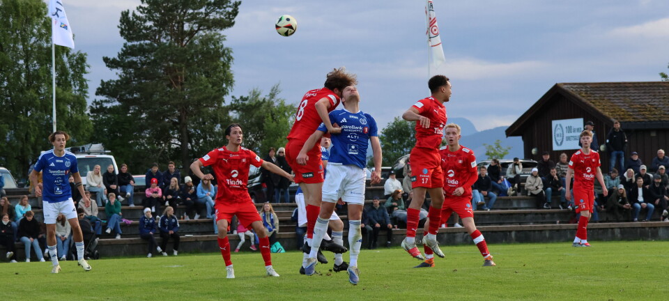 Spillere i røde og blå drakter kjemper om en høy ball på en fotballbane.