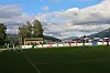 Fotballbane med fotballspillere, mål, reklameskilt og fjell i bakgrunnen under delvis skyet himmel.