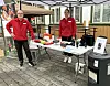 To personer ved et bord under et telt med brosjyrer, utstyr og en varmepumpe på en uteplass.
