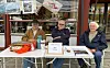 Tre menn sitter ved et bord utenfor en kafé med aviser, papirer og en termos.