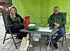 To personer sitter ved et bord under et grønt telt med drikke og papirer.