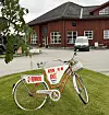 Rød sykkel med kurv står på gresset foran et rødt trebygg.