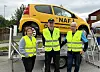 Tre personer i refleksvester står foran en gul bil med NAF-logo som er løftet opp.