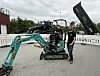 En mann står ved og en mann sitter inne i en liten grønn minigraver foran en traktor og en henger på et utstillingsområde.
