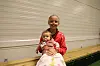 Barn i rød jakke sitter på en benk og holder en baby i en innendørs hall.