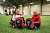 To jenter sitter på grønt kunstgress i en innendørshall med røde og svarte fotballbager foran seg.