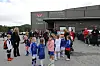Barn står samlet utenfor en sportsarena med voksne og flagg i bakgrunnen.