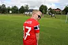 Ung spiller i rød fotballdrakt står på en gressbane med mål og spillere i bakgrunnen.