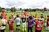 En gruppe barn og ungdommer står samlet på en fotballbane med trenere i bakgrunnen.