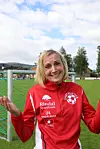 Smilende kvinne i rød sportsjakke står ved et fotballmål på en grønn fotballbane.