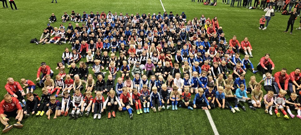 Stor gruppe mennesker sitter og står samlet på en grønn fotballbane innendørs.