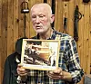 En mann holder et innrammet fotografi foran en trevegg med hengende verktøy.