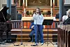 En sanger står ved mikrofoner foran alteret i en kirke mens publikum sitter på benker.