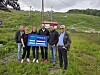 En gruppe mennesker står ved en stolheis og holder opp et blått skilt i et grønt fjellområde.