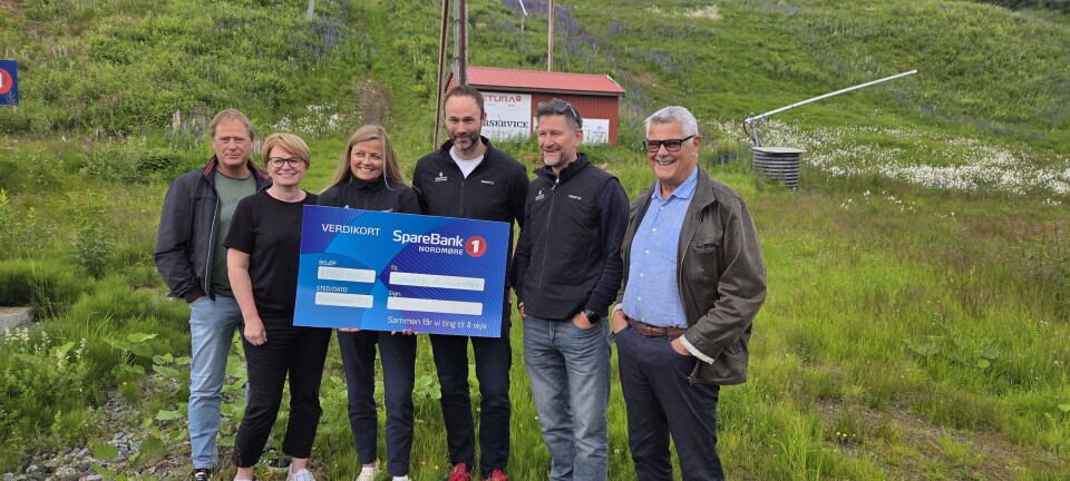 En gruppe mennesker står ved en stolheis og holder opp et blått skilt i et grønt fjellområde.