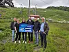 En gruppe mennesker står ved en stolheis og holder opp et blått skilt i et grønt fjellområde.