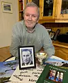 Eldre mann sitter ved et bord med et innrammet foto, en pokal og bøker i en stue.